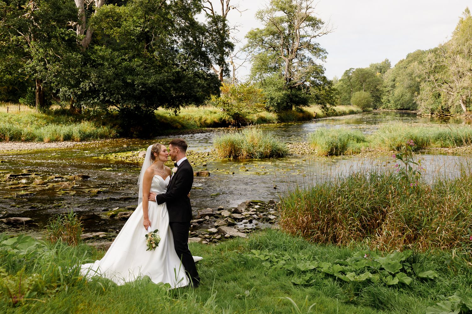 A couple shares a romantic moment by a tranquil pond surrounded by lush greenery and natural scenery.