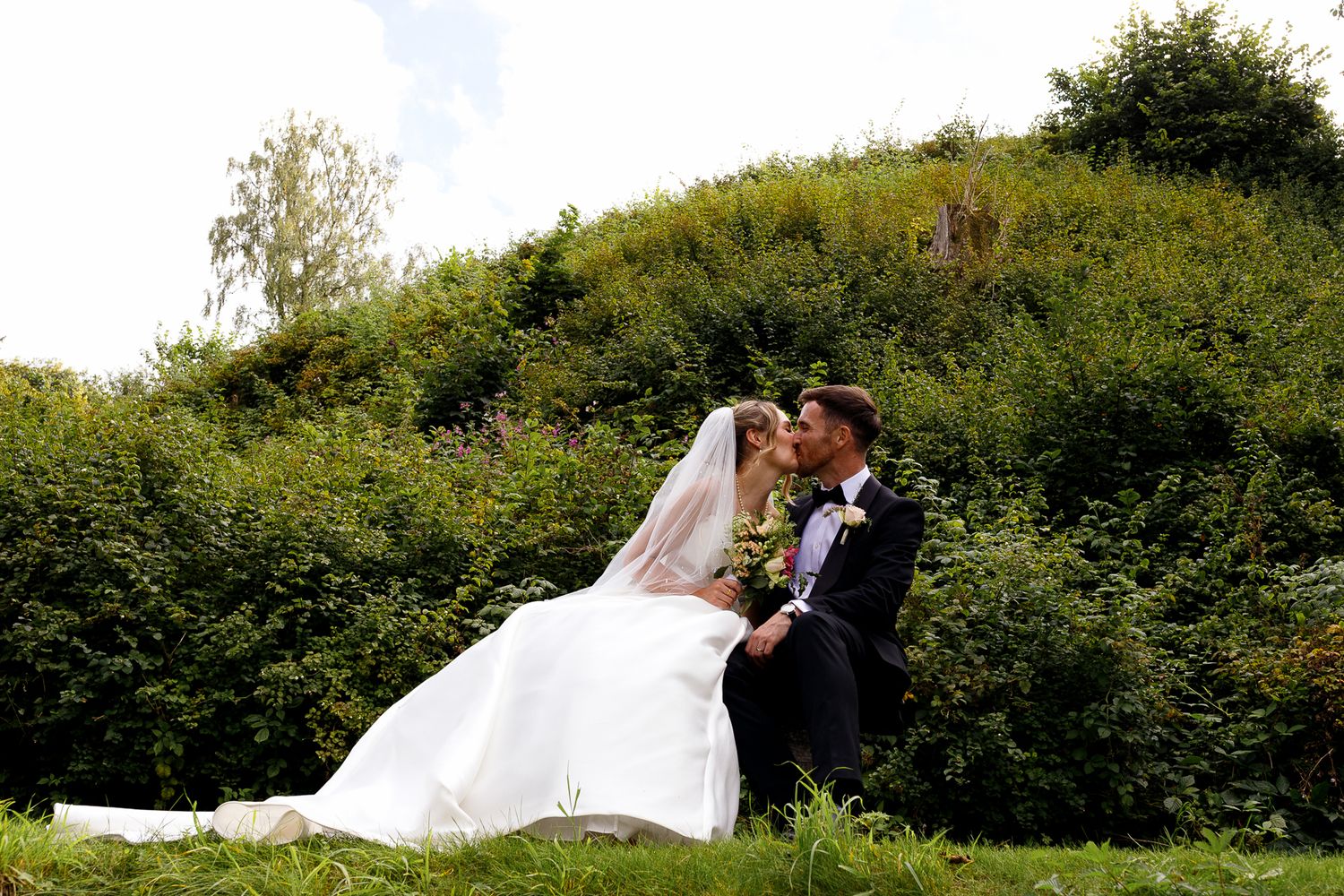 A romantic wedding couple shares a tender moment in a lush green field at sunset.