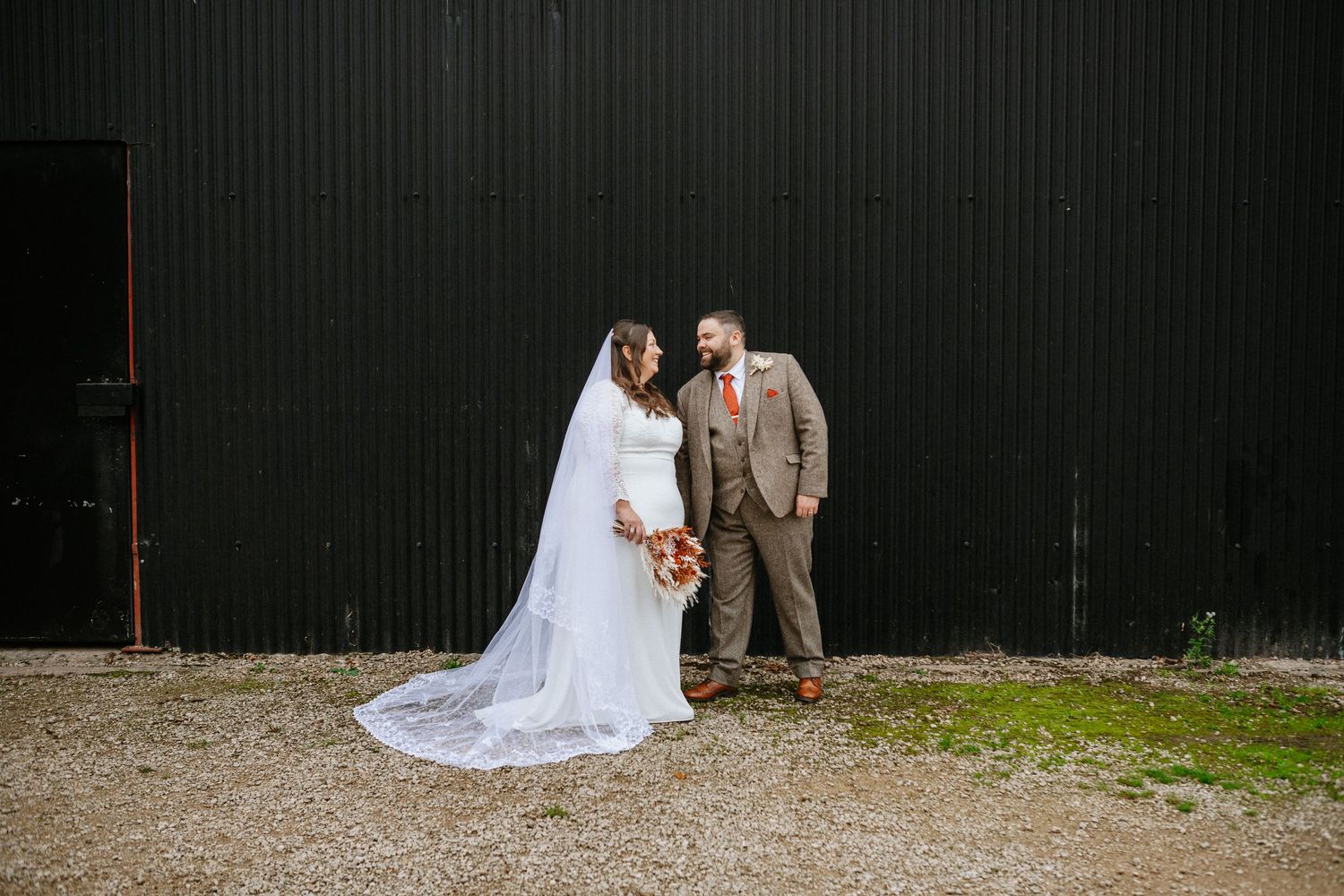 A couple in wedding attire poses together in front of a rustic green barn wall at Donington Park Farmhouse wedding venue.