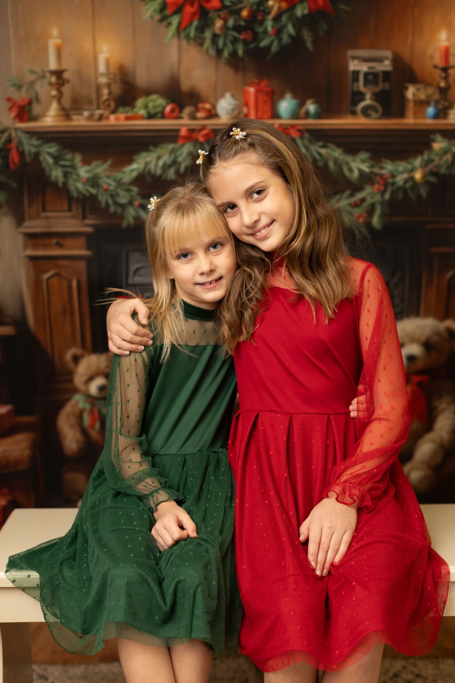 Two children in festive holiday dresses pose together against a decorated mantel background with Christmas decorations.