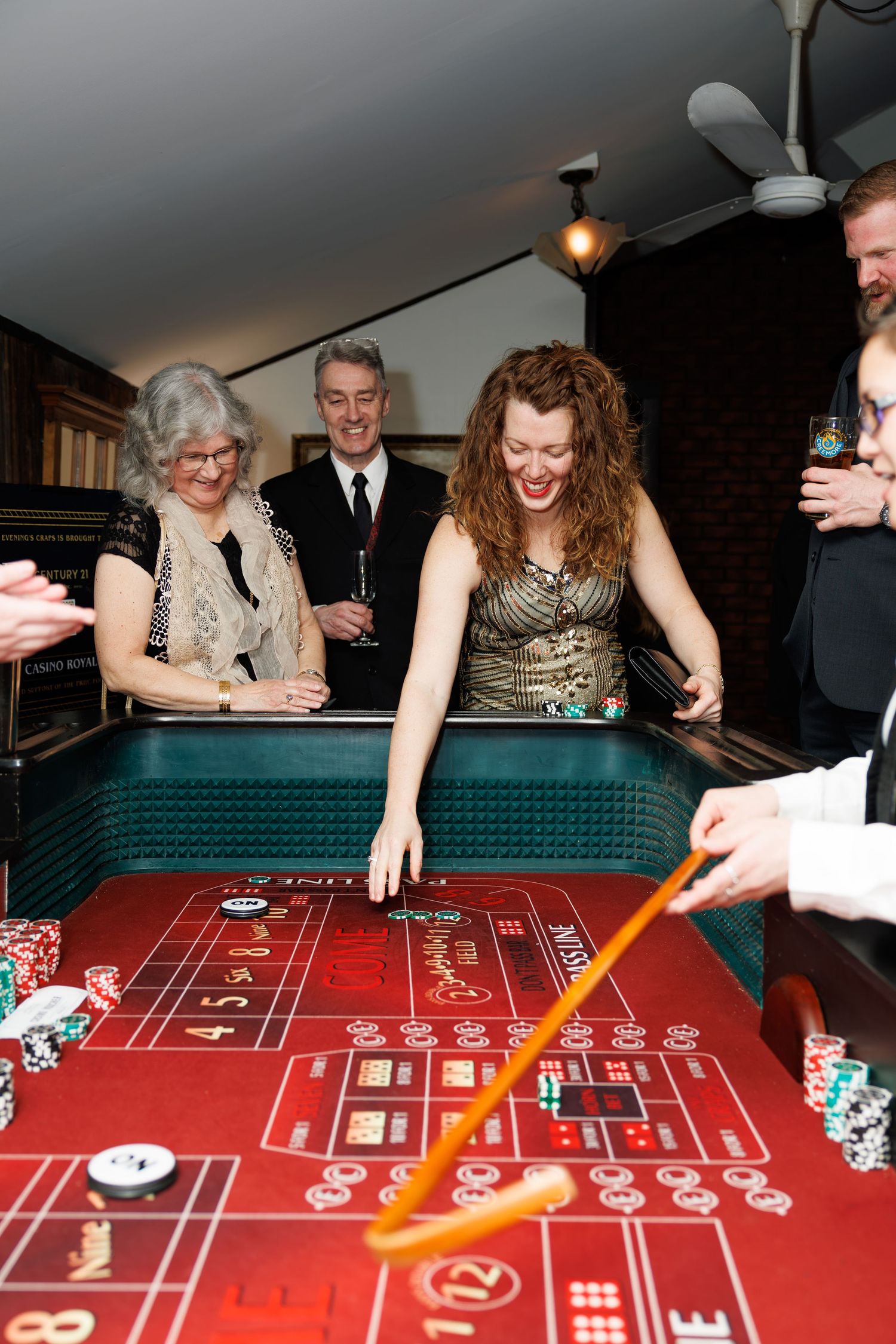 Group of people laughing and playing craps at a casino table with colorful poker chips during a formal event.