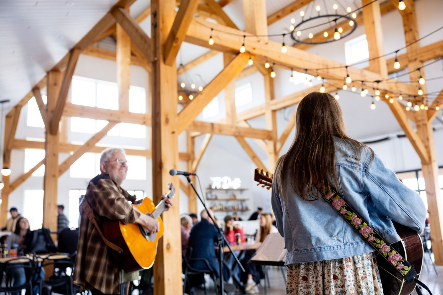 Two musicians perform acoustic guitars in a rustic timber-frame venue with string lights during a live event.