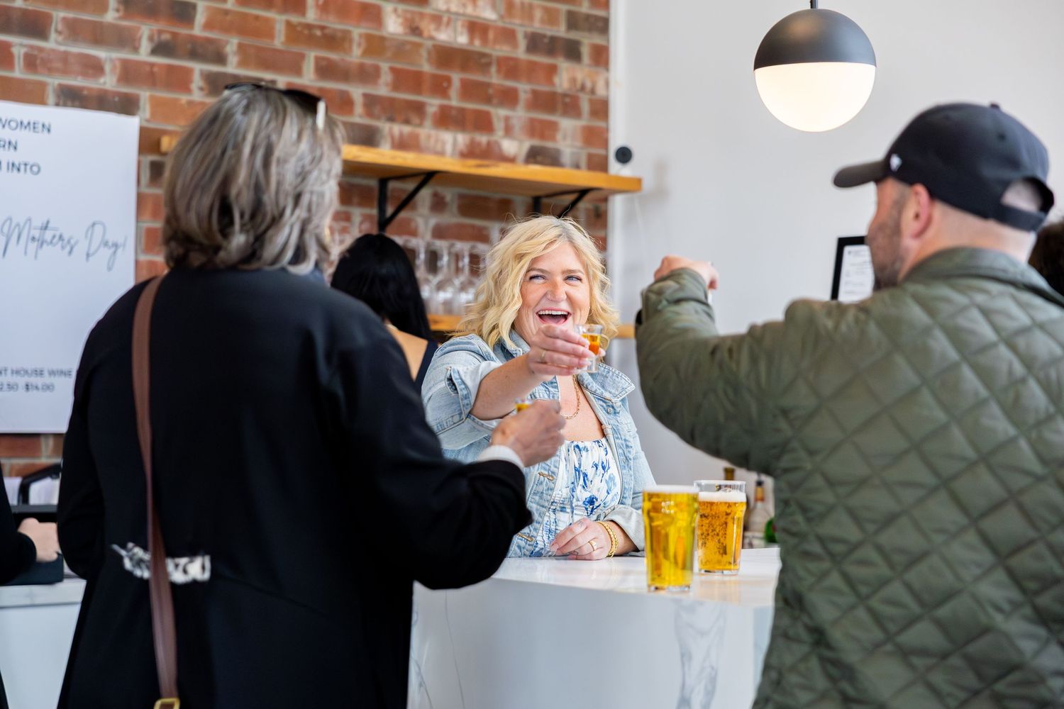 People socializing at an indoor event, laughing and enjoying drinks at a white counter with brick walls.