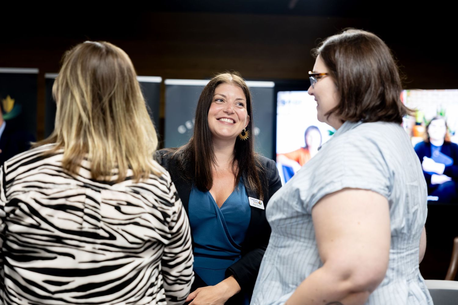 Three women engaged in conversation at a professional networking event, smiling in front of digital displays.