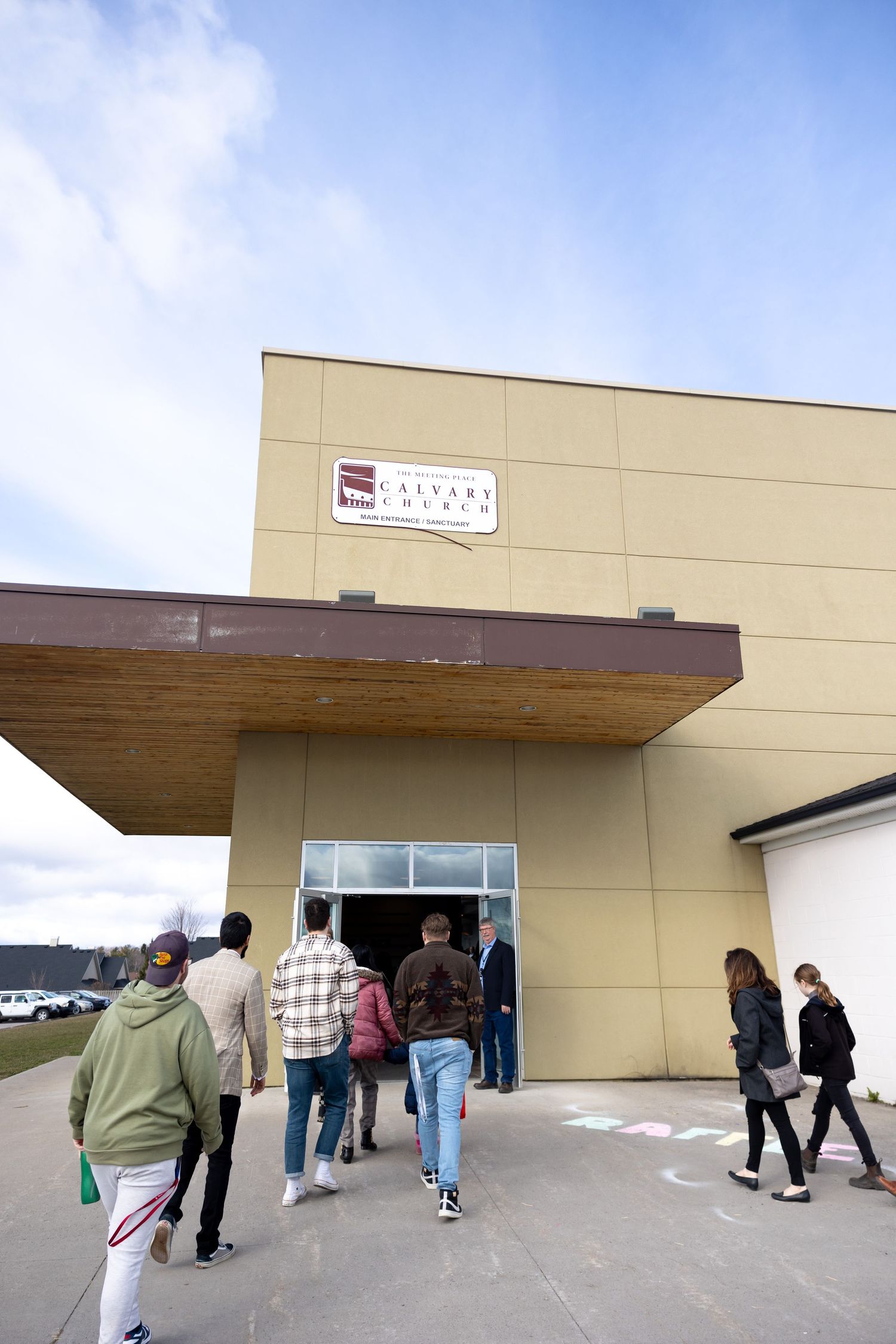 People wait in line outside a beige commercial building with glass doors on a cloudy day.