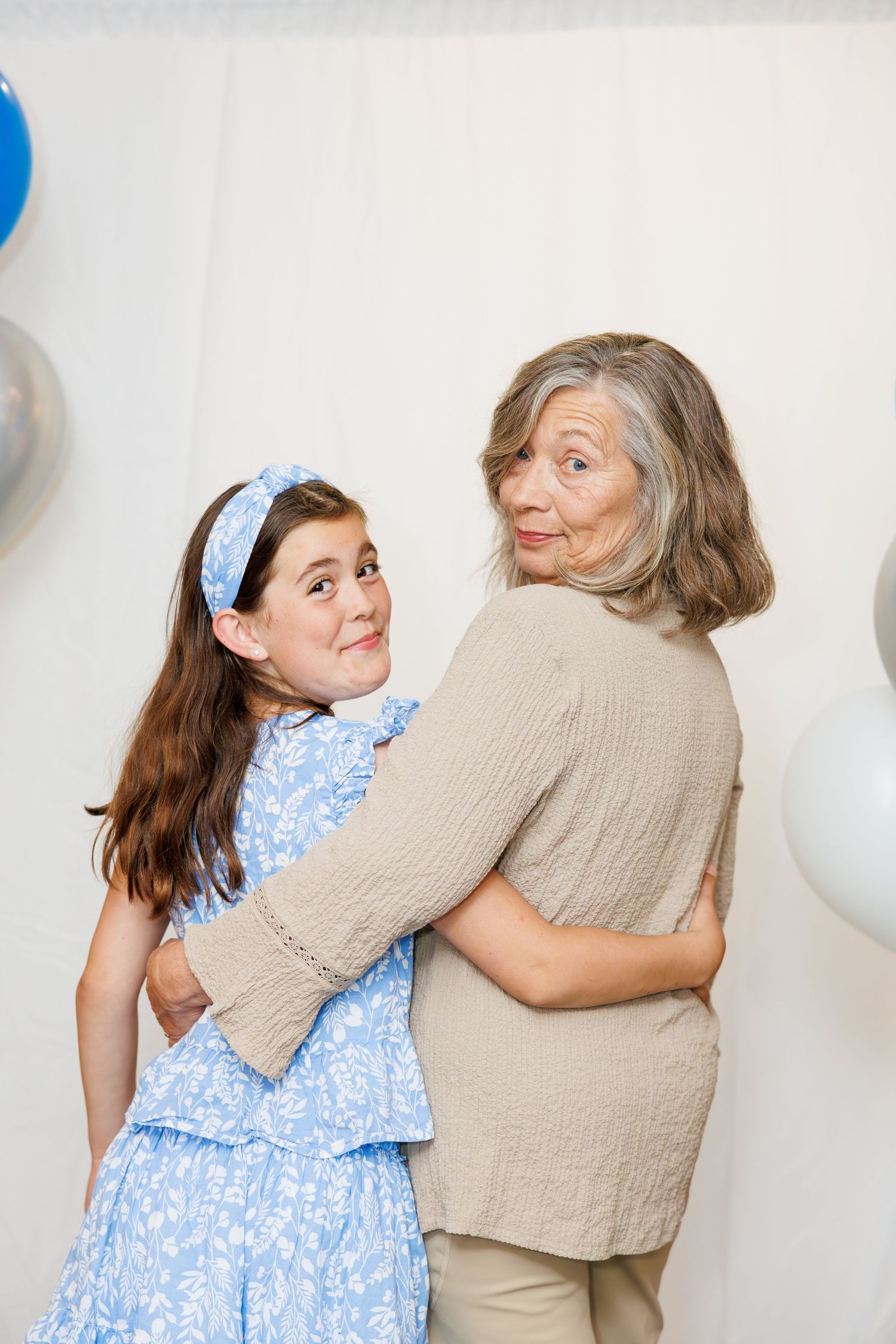 Two people embrace while posing against a white wall with blue balloons in a playful portrait setting.
