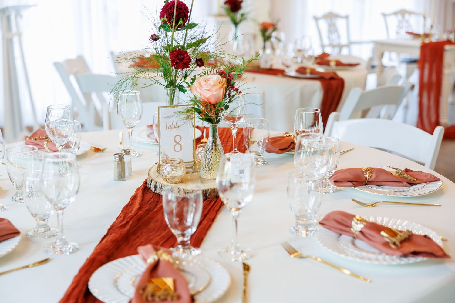 Elegant wedding reception table settings with red and peach floral centerpieces and white chairs in a bright venue.