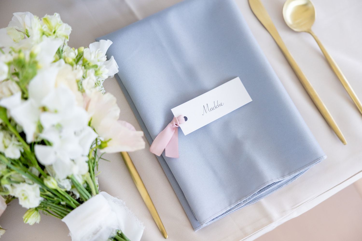 White flowers and gold utensils arranged with gray napkins on a light surface create an elegant table setting.
