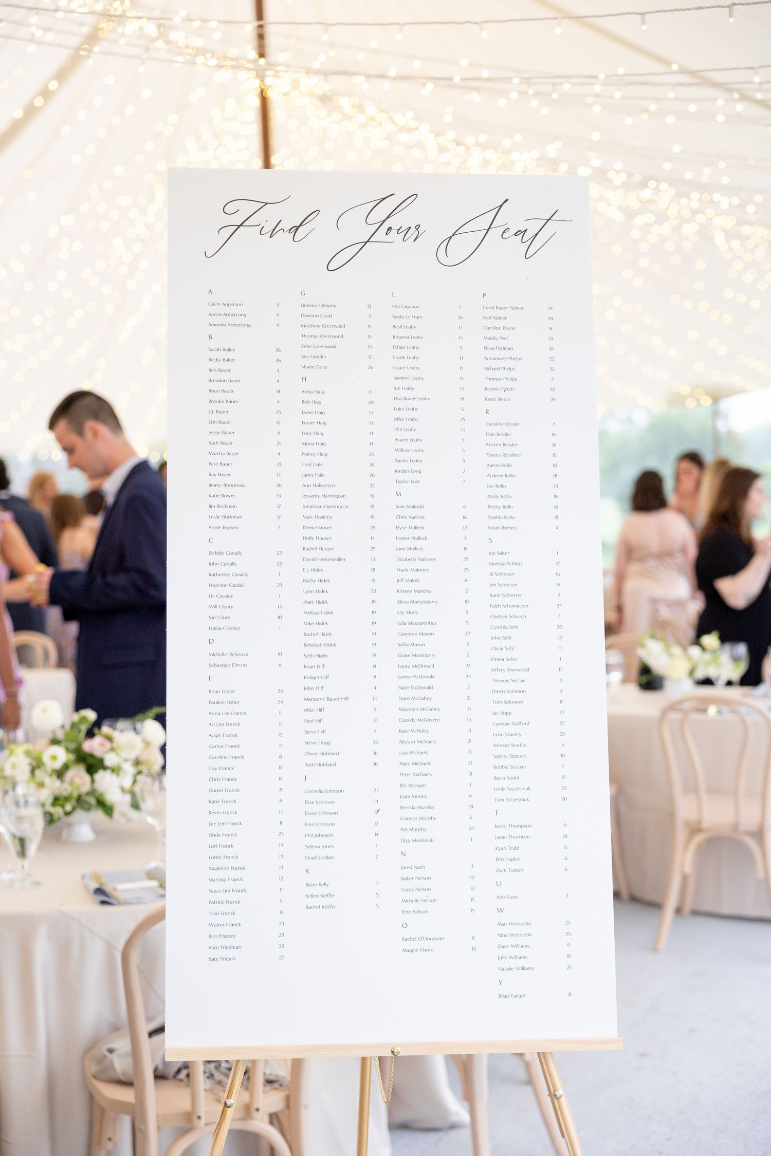 Elegant wedding seating chart display with white paper lists mounted on wooden easels in a light-filled venue.