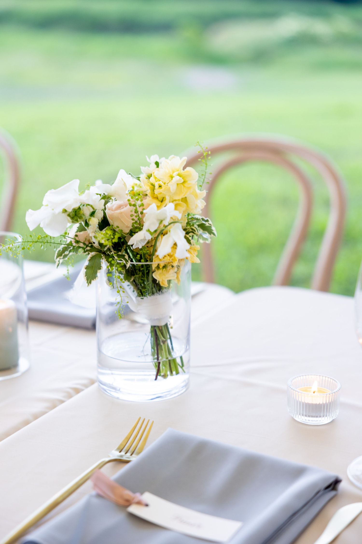 Elegant outdoor wedding table setting with white linens, gold cutlery and delicate floral centerpieces in soft daylight.