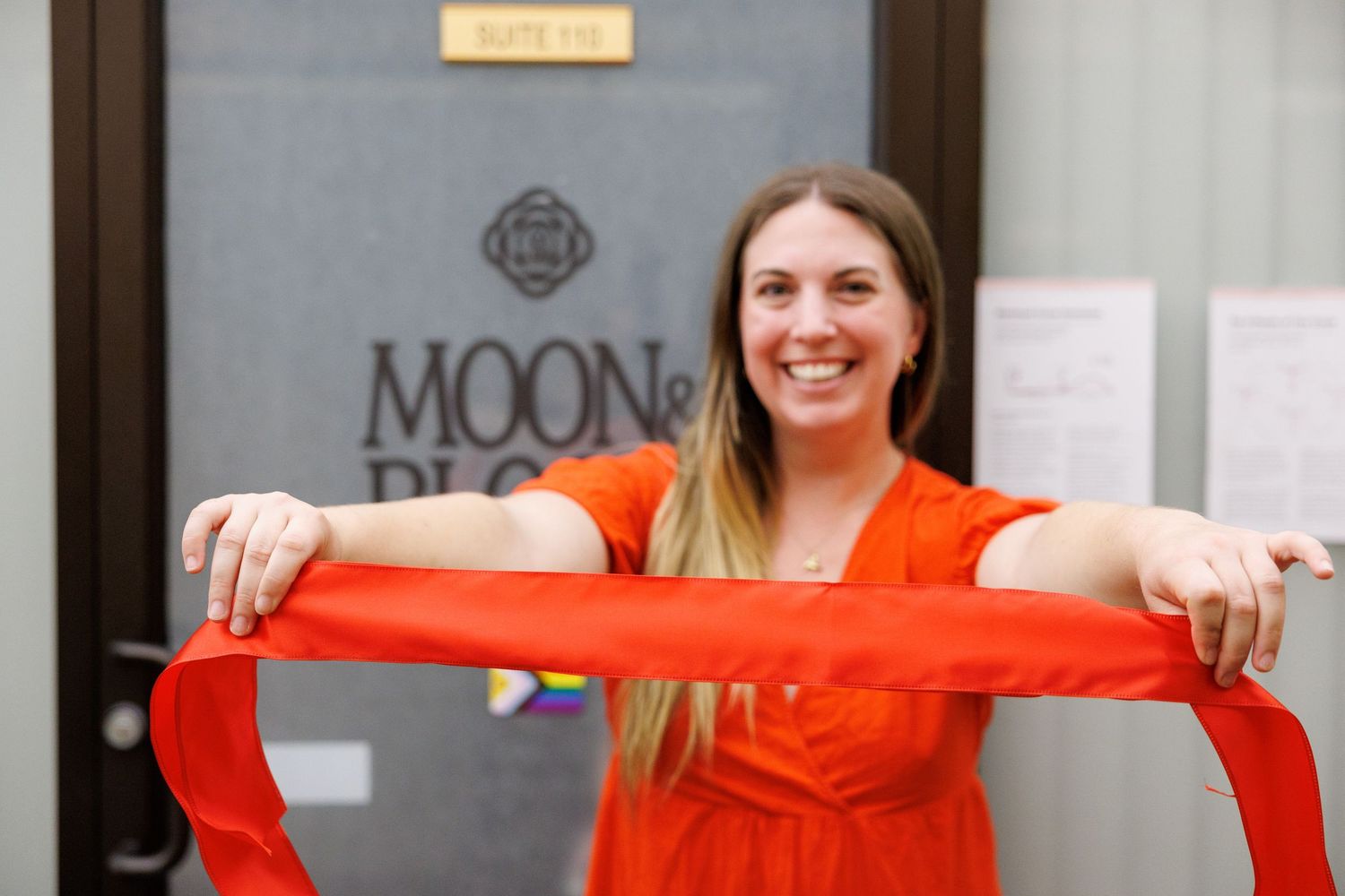 A person in a bright orange shirt holds up a red ribbon in front of a storefront window.