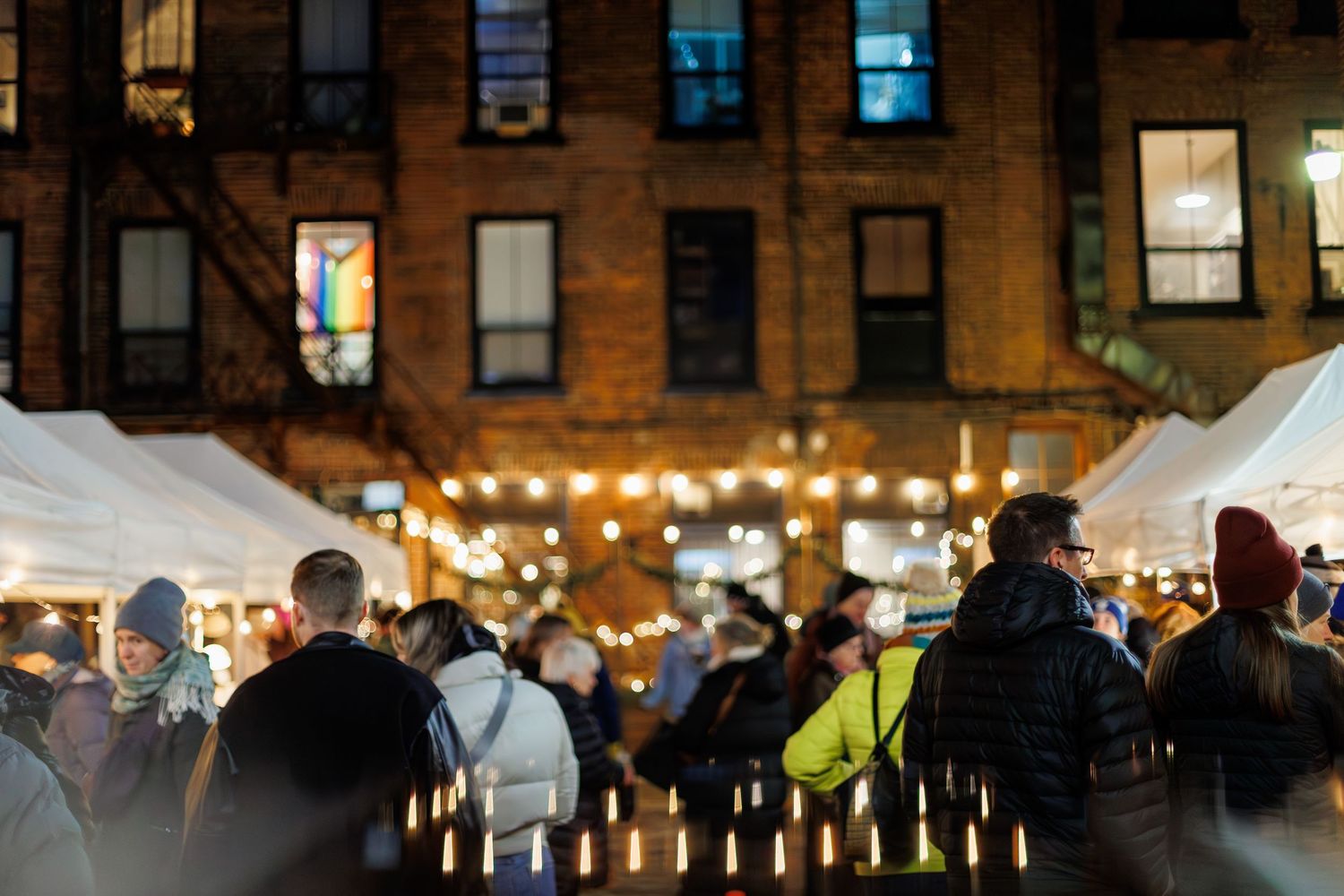Crowded outdoor holiday market at night with string lights and white tents in front of a brick building.