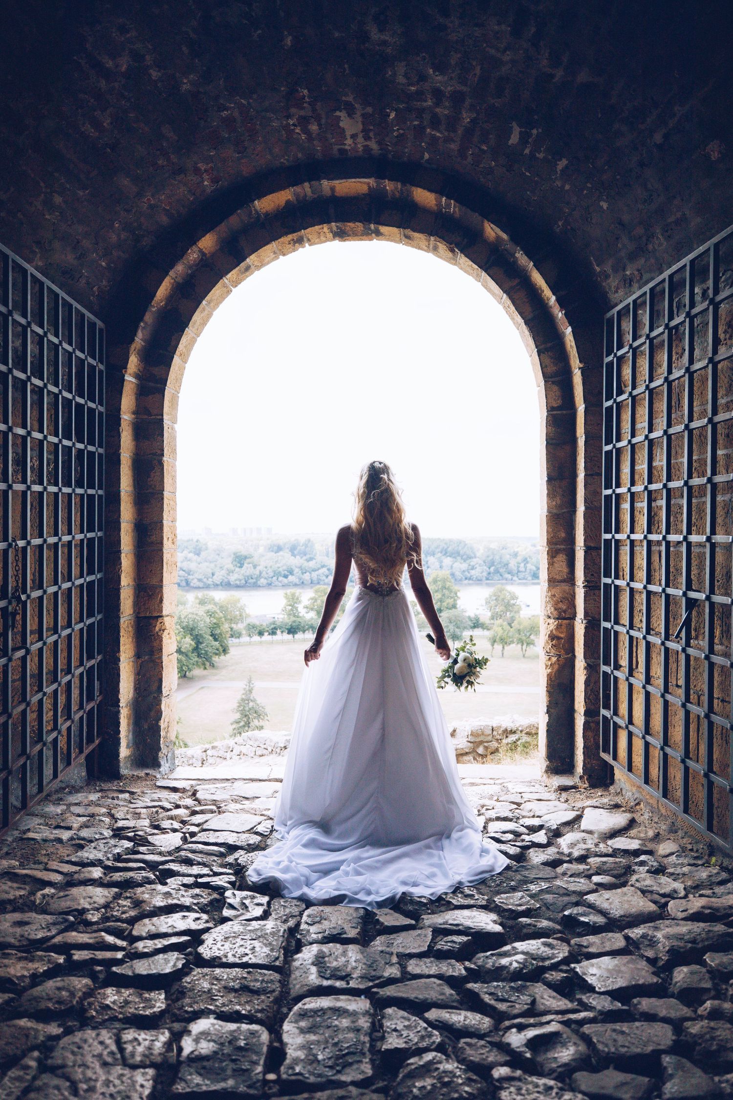 A figure in a white wedding dress stands silhouetted in a stone archway overlooking a scenic landscape.