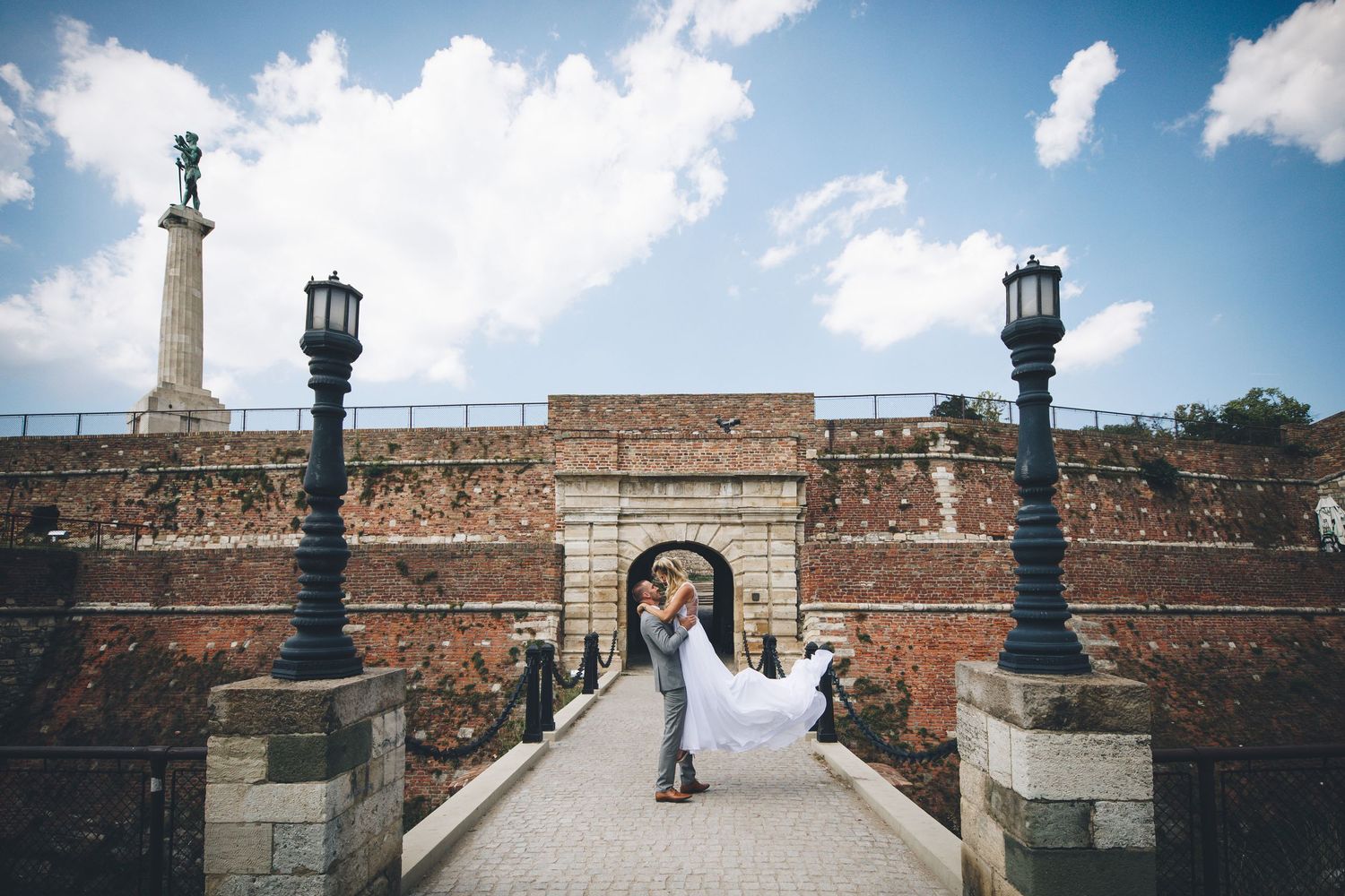 A panoramic wedding photo sequence showing a couple on a brick terrace with ornate lampposts against a blue cloudy sky.