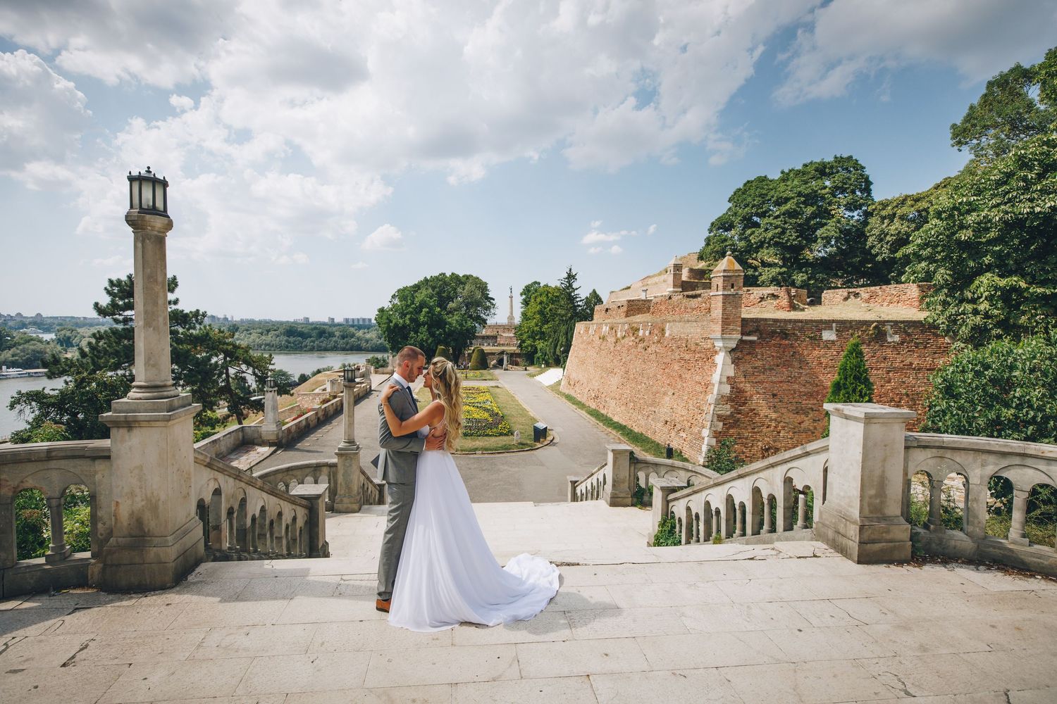 A romantic couple embraces on stone steps overlooking a scenic European city with historic fortress walls and greenery.