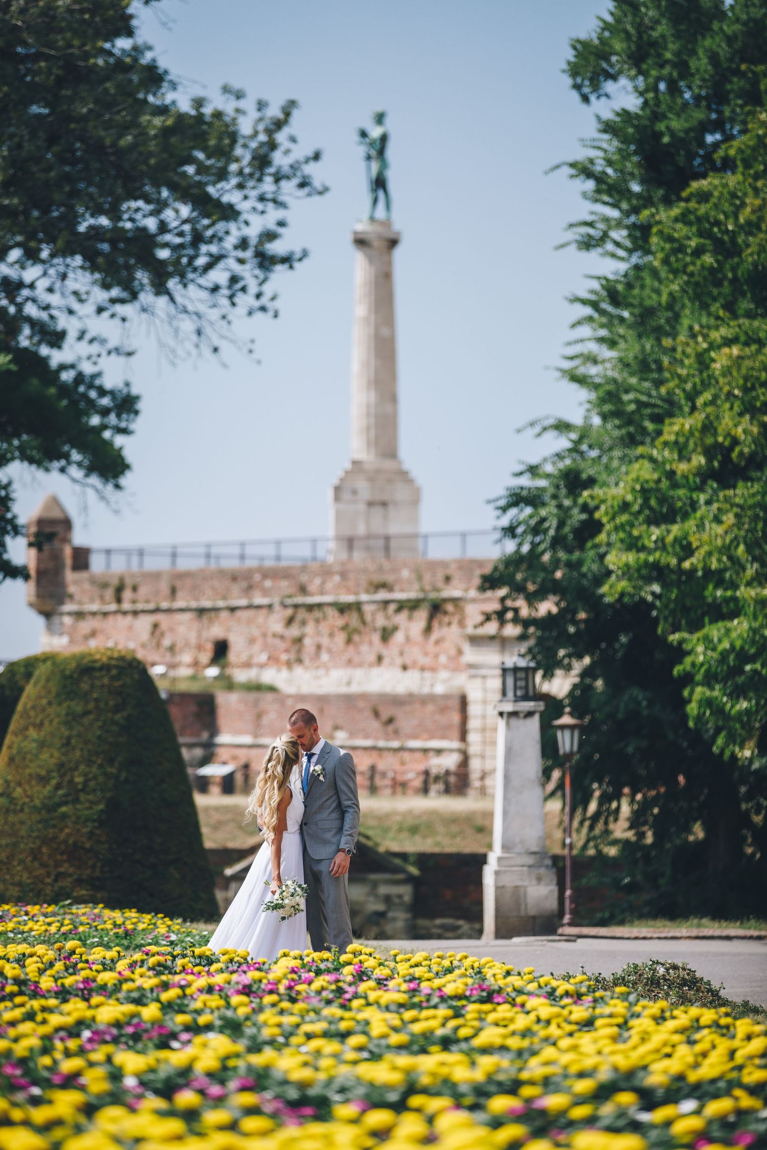 A romantic couple shares a tender moment in a garden with vibrant yellow flowers and a stone monument in the background.