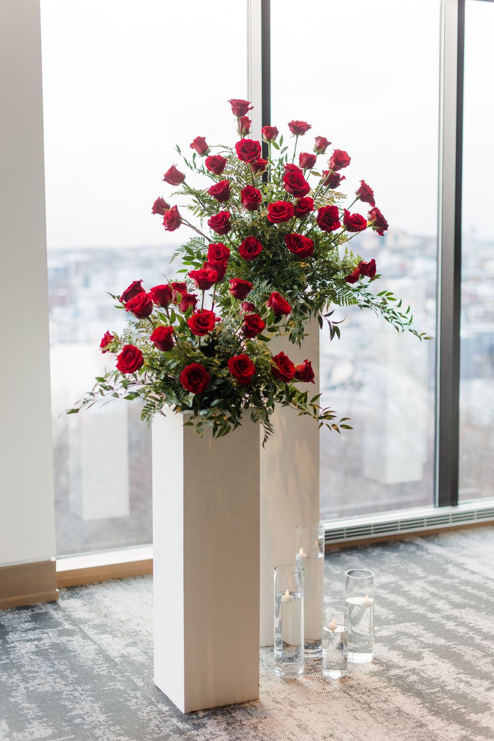 Elegant red rose floral arrangement in tall white pedestal with greenery against window overlooking city view.