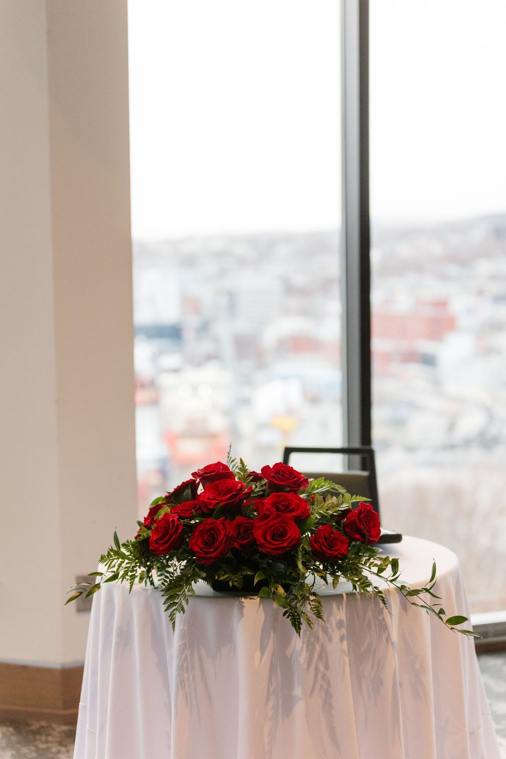 Red rose floral arrangements adorn white tablecloths with a cityscape view through large windows in the background.