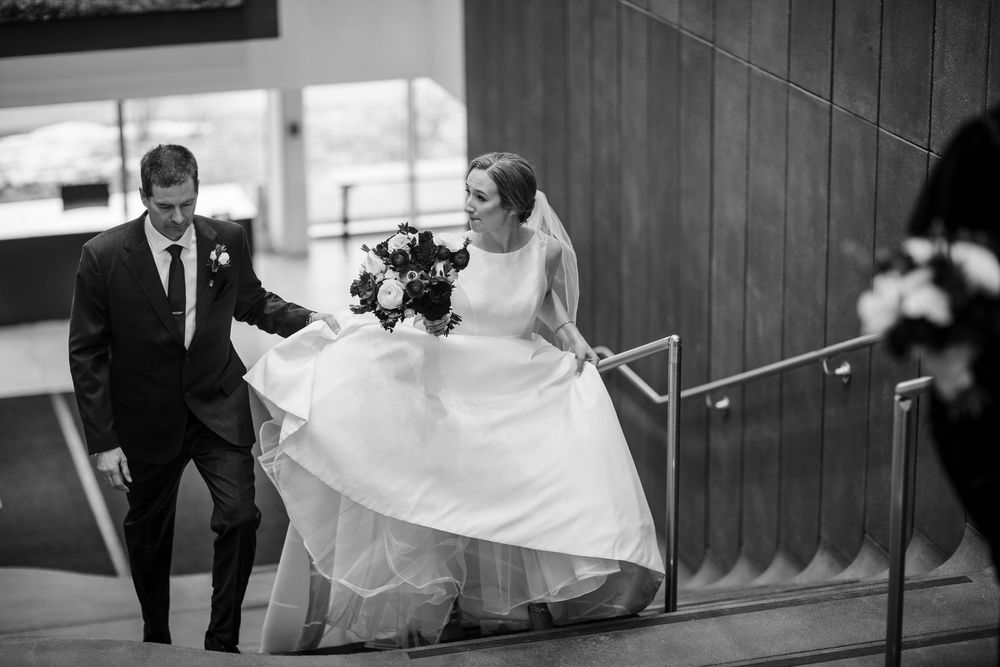 A bride in a flowing white gown descends a staircase with assistance while holding a floral bouquet in black and white.