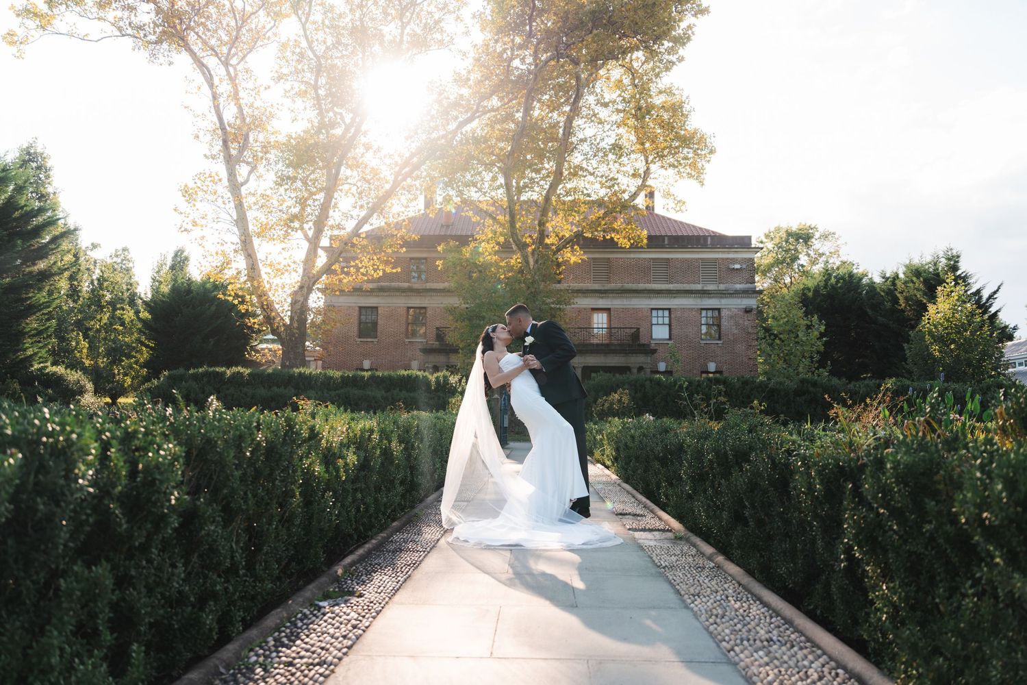 Wedding couple walks along a scenic garden path surrounded by lush greenery and buildings during golden hour.