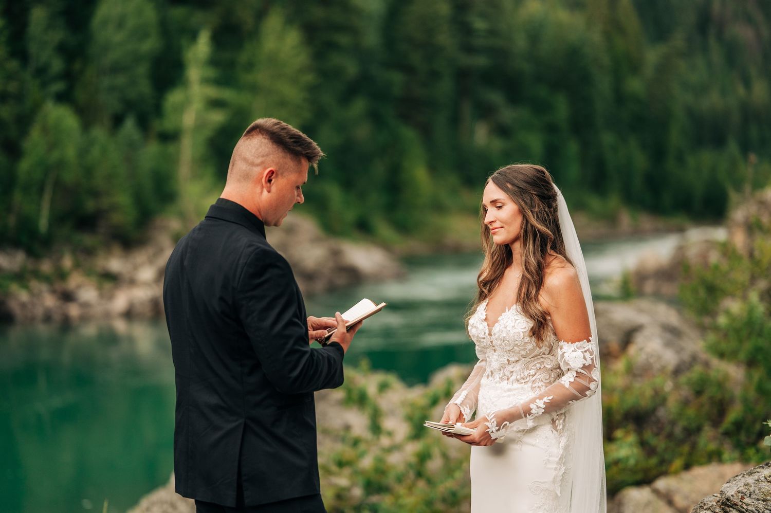 Couple exchanges vows during outdoor wedding ceremony beside a serene mountain river in a lush forest setting.