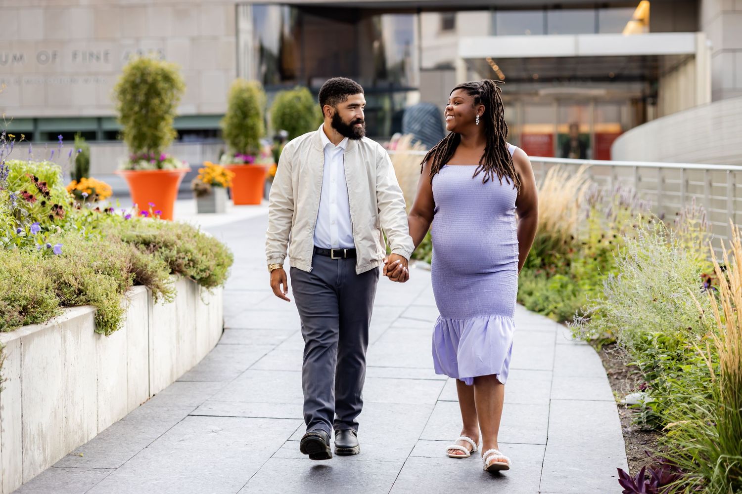 A couple walks together through a garden path on a sunny day wearing purple and gray attire.