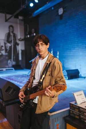 A musician performs with an electric guitar in a blue-lit venue wearing a tan jacket and white t-shirt.