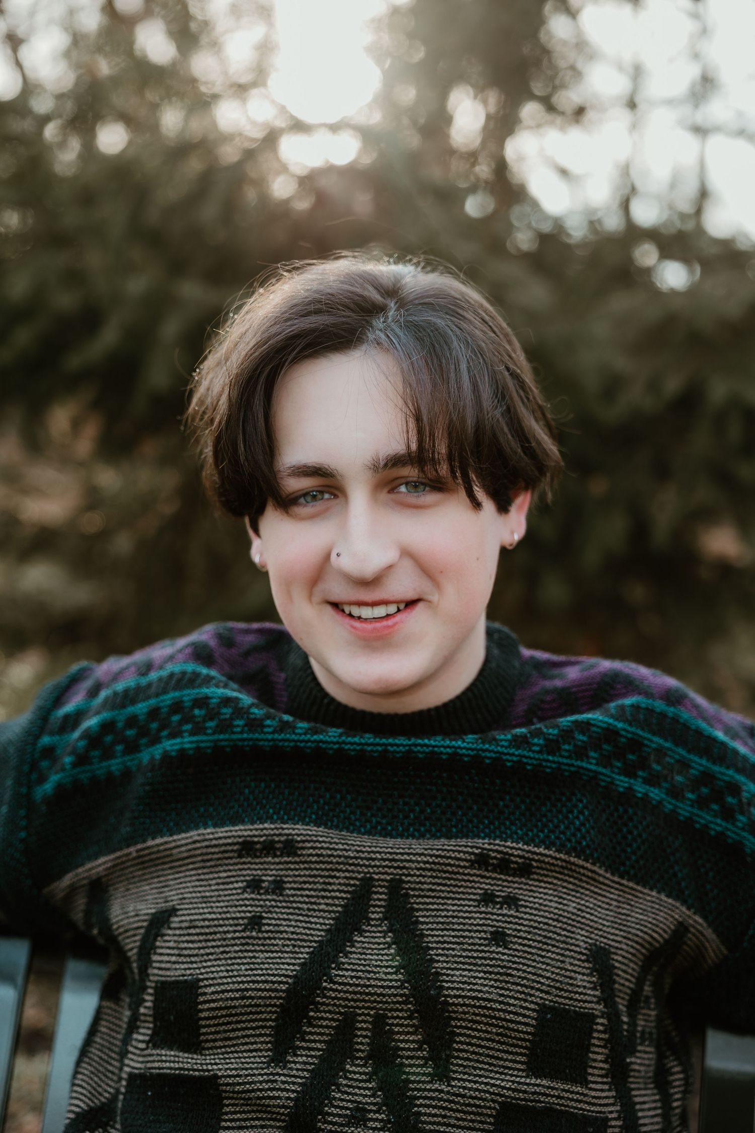 A person wearing a dark knit sweater smiles warmly outdoors during golden hour lighting.