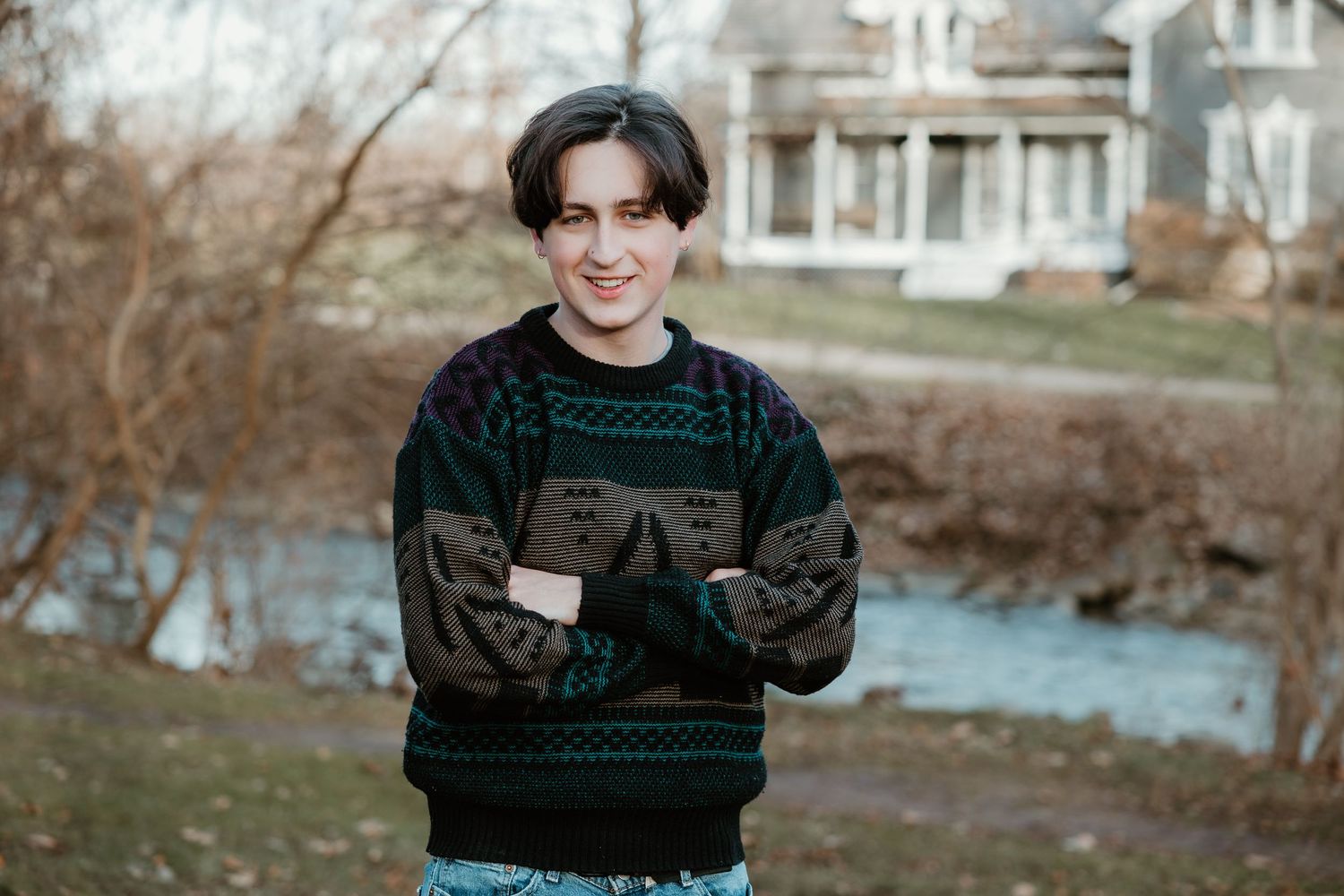 A person wearing a dark patterned sweater smiles while standing outdoors near a pond and house on a winter day.