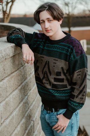 Person wearing a dark knit sweater with teal accents and geometric patterns poses against a light colored wall.