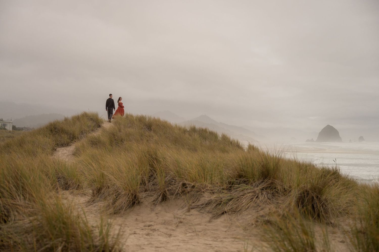 A young couple walks through a grassy coastal dune on an overcast day near Cannon Beach, OR. Haystack rock is pictured in the distance.