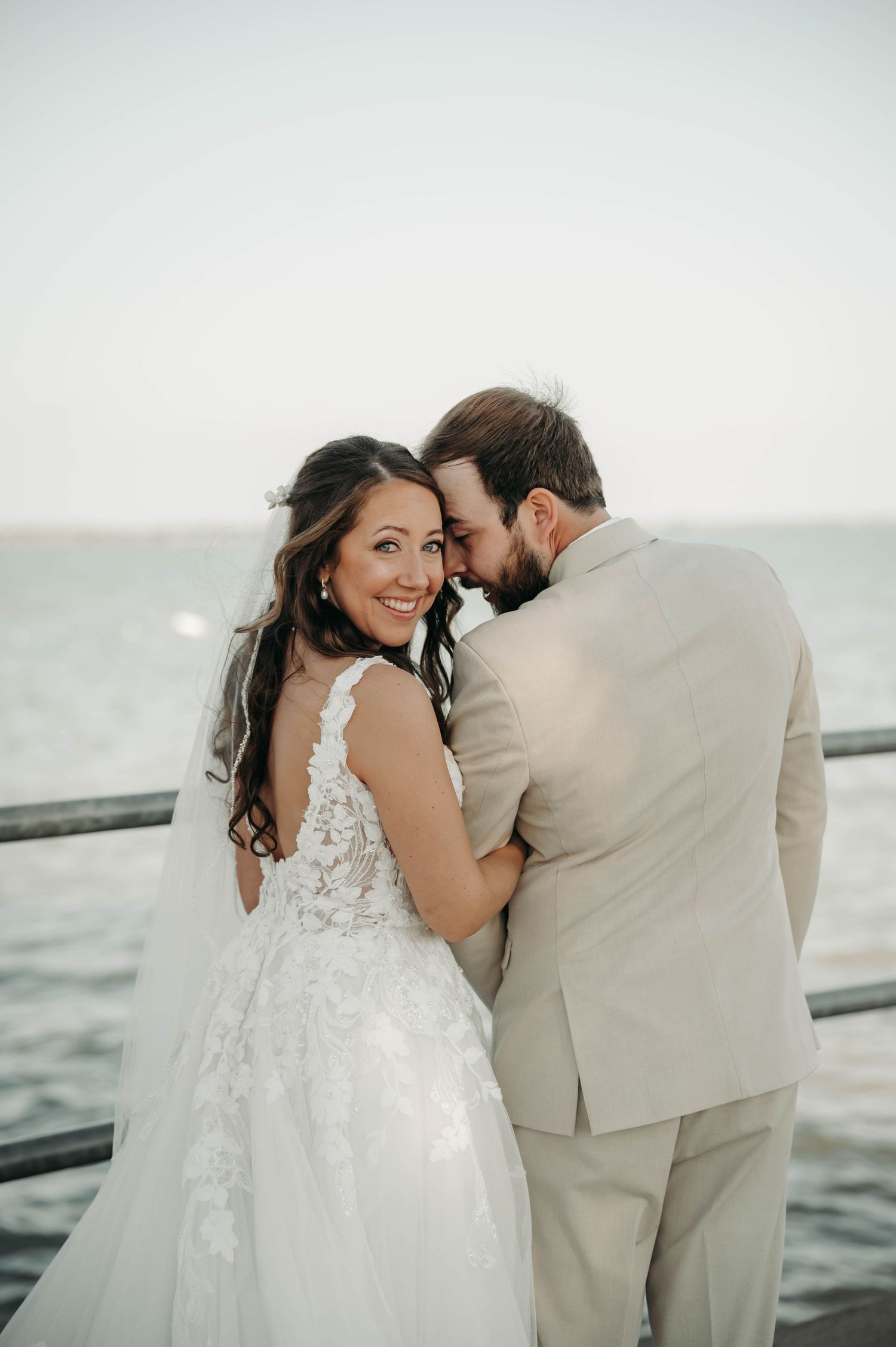 A smiling bride in a lace gown embraces her groom in a tan suit on a waterfront pier.