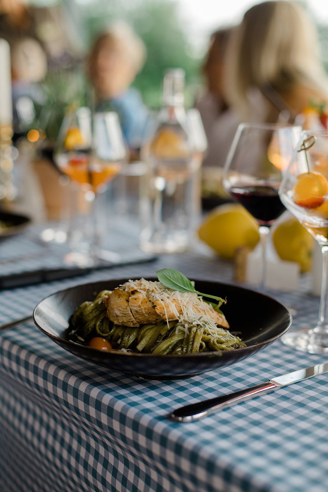 A series of plated pasta dishes with garnish on black plates sit atop blue and white checkered tablecloths.