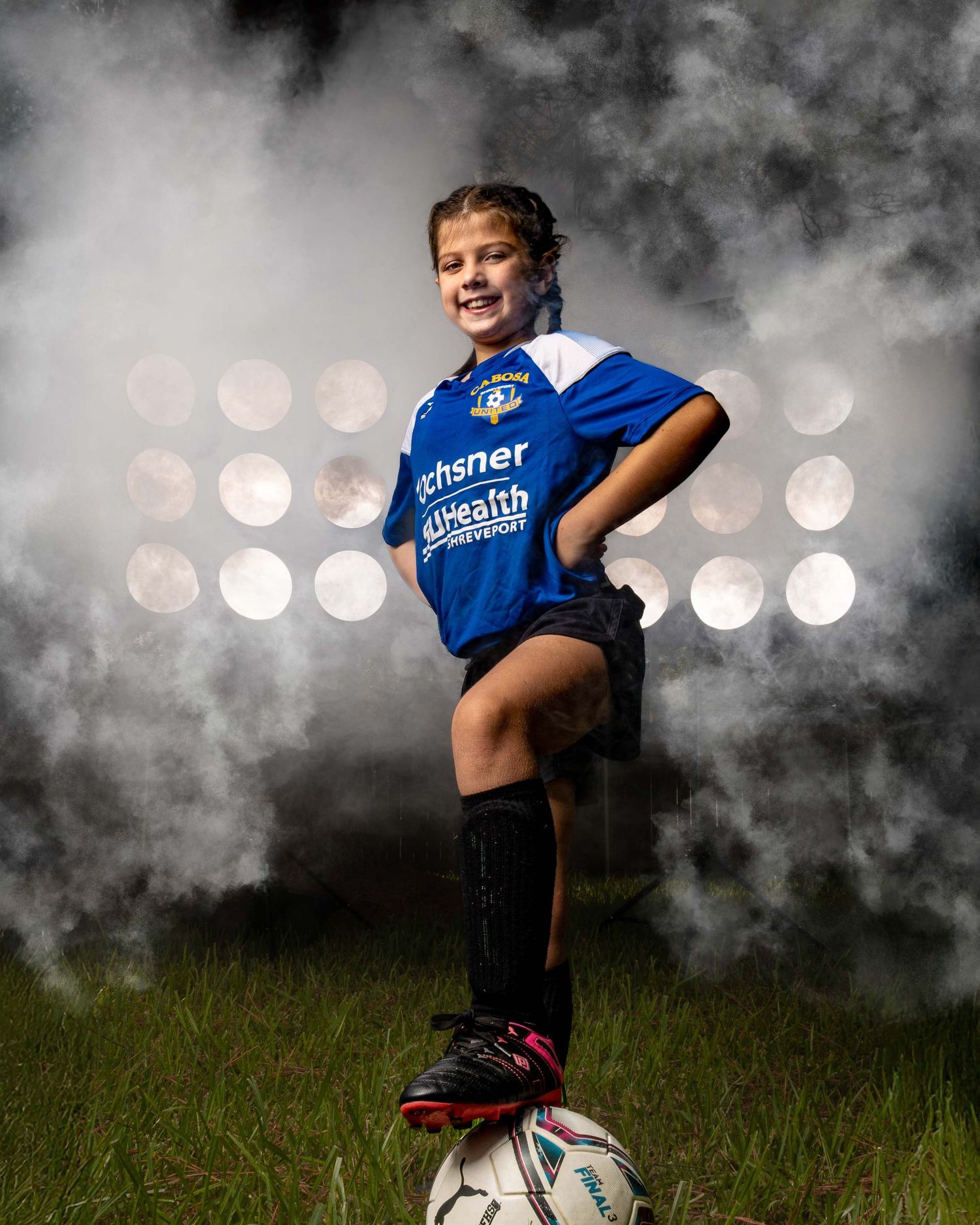 Young soccer player in blue jersey poses confidently with foot on ball against dramatic backlit background with bokeh lights.