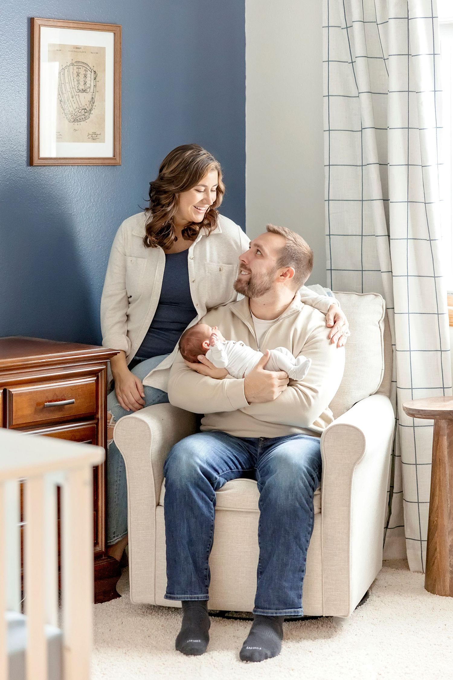 A Garner, Iowa couple sits together on a cream-colored armchair in a cozy nursery with natural lighting during their newborn session.