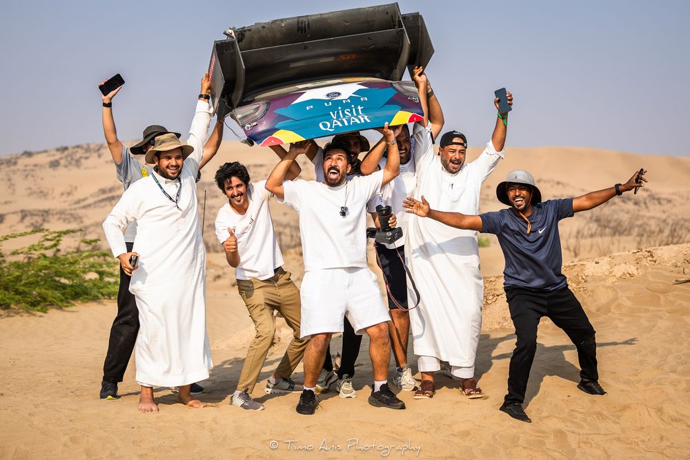 A group of people celebrating in traditional Middle Eastern dress while sandboarding in a desert landscape.