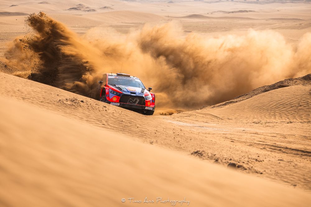 A red rally car kicks up sand while racing through desert dunes during an off-road competition.