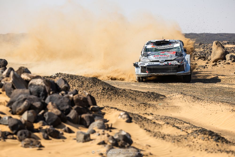 Rally car kicks up dust while racing through desert terrain during off-road competition.