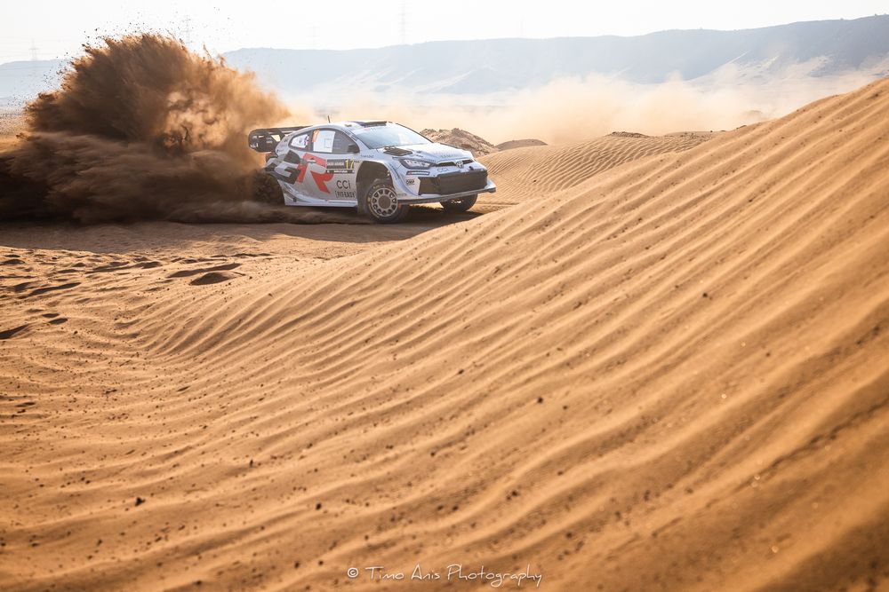Off-road vehicle drives through golden sand dunes in desert landscape during sunset.