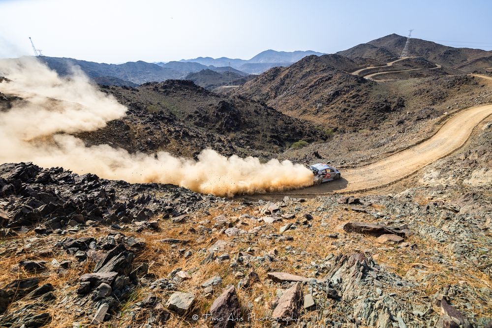 Aerial panoramic view of rugged mountain terrain with rocky slopes and winding paths under a bright sky.