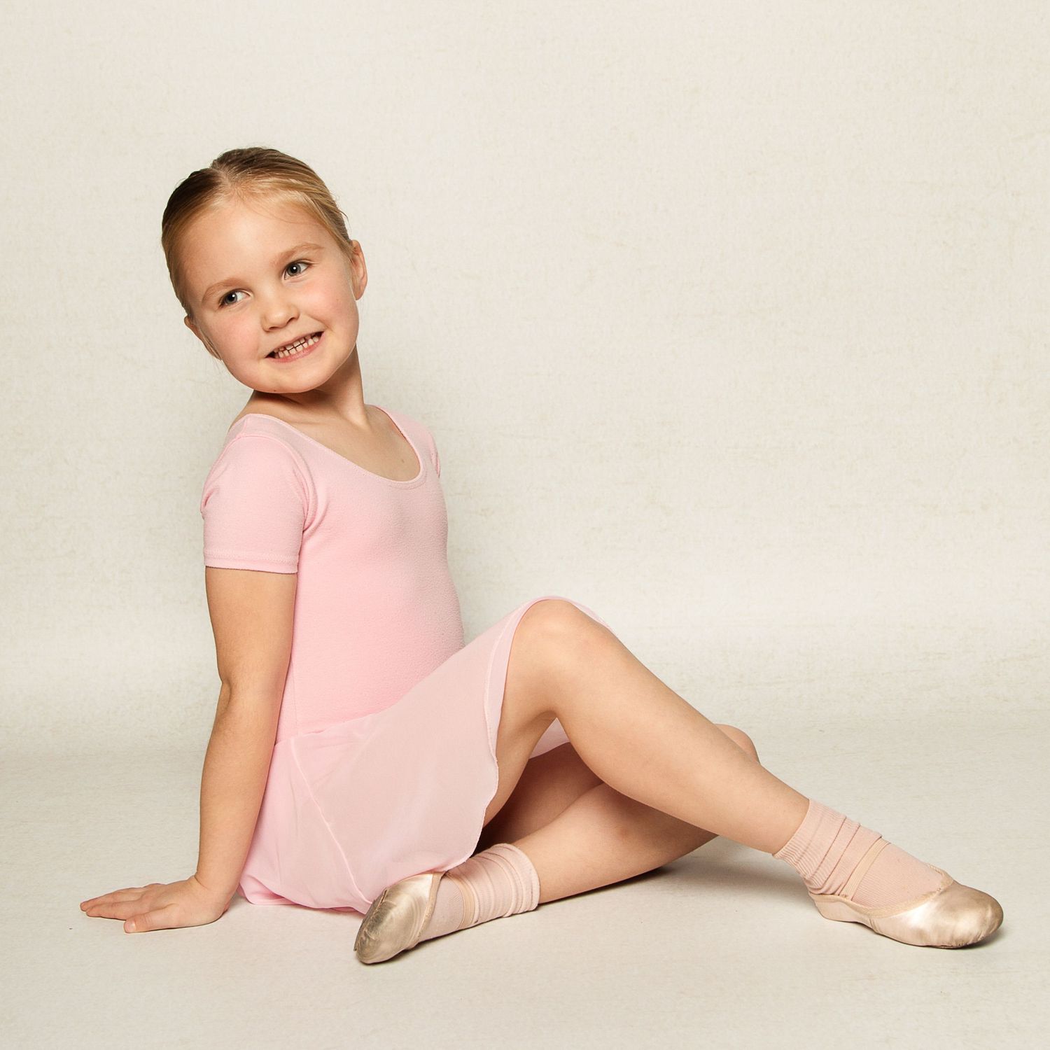 Young girl in her ballet outfit sitting on the floor in a ballet pose smiling.  She is on a white background in a studio location.
