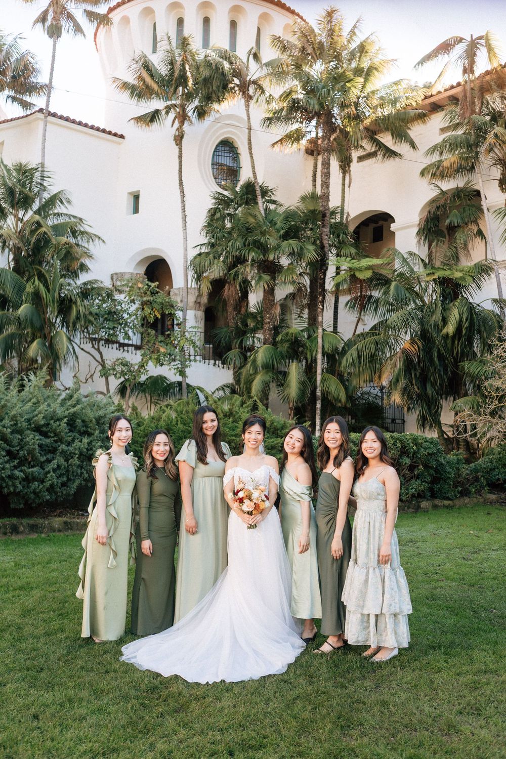 Bridesmaids in sage green dresses pose in front of palm trees at the Santa Barbara Courthouse