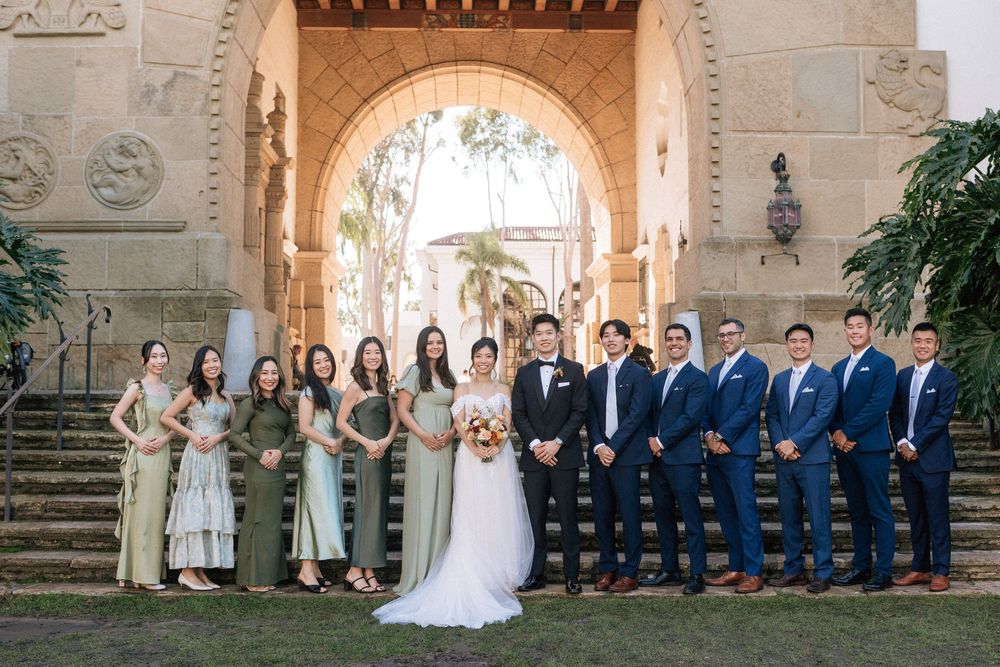 A large wedding party with bridesmaids in sage green dresses and groomsmen in suits posing on the staircase at Santa Barbara Courthouse