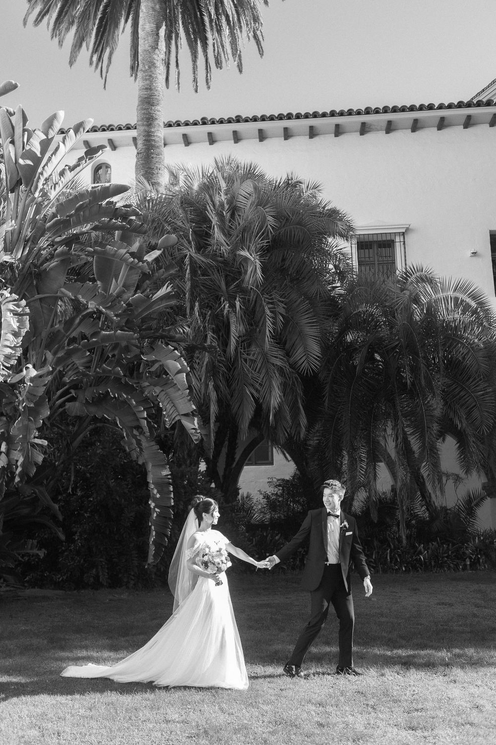 A romantic black and white photo of a bride and groom walking hand in hand along the Santa Barbara Courthouse Sunken Gardens lawn.