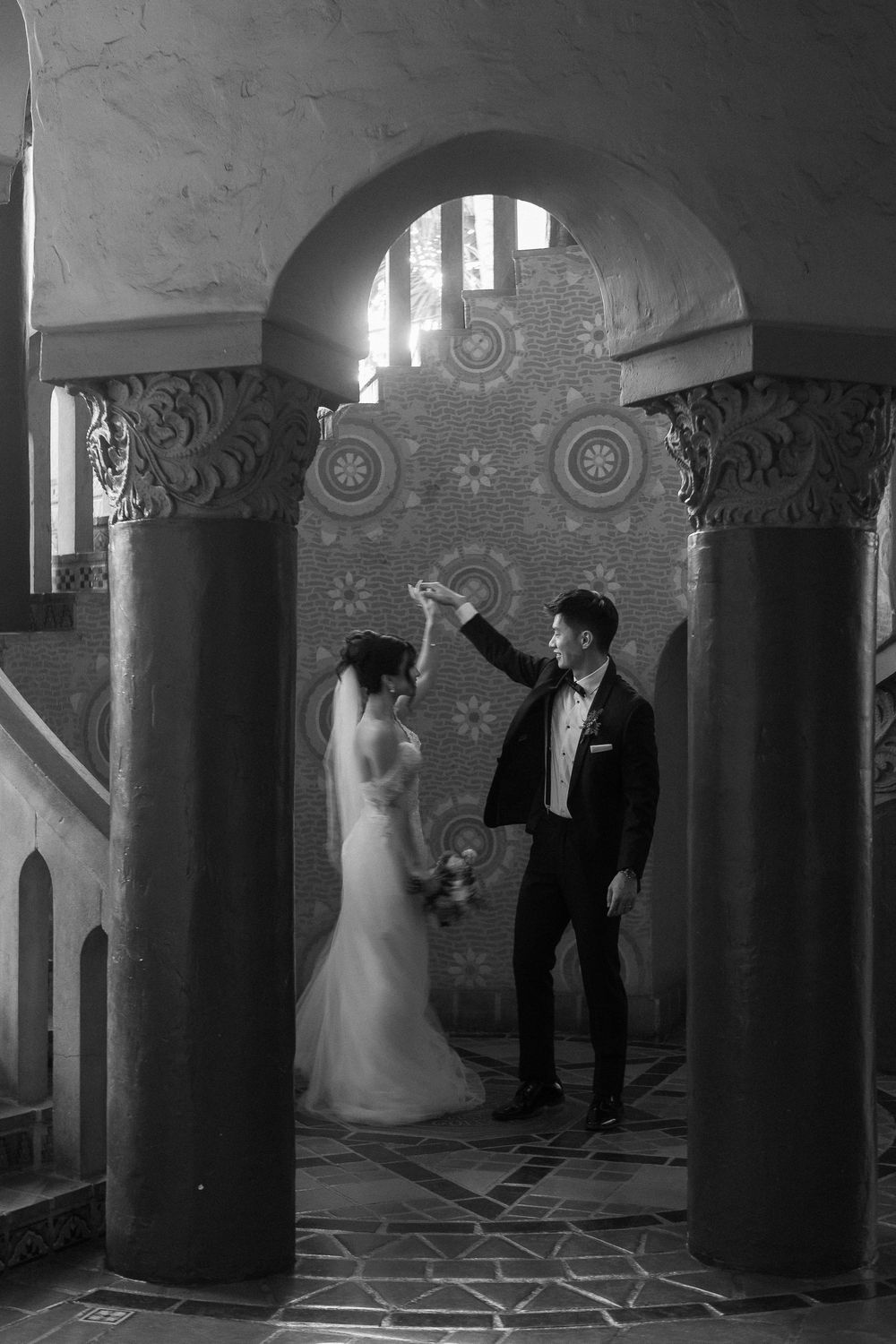 A black and white photo of a groom twirling a bride in a gown under the staircase of the Santa Barbara Courthouse.