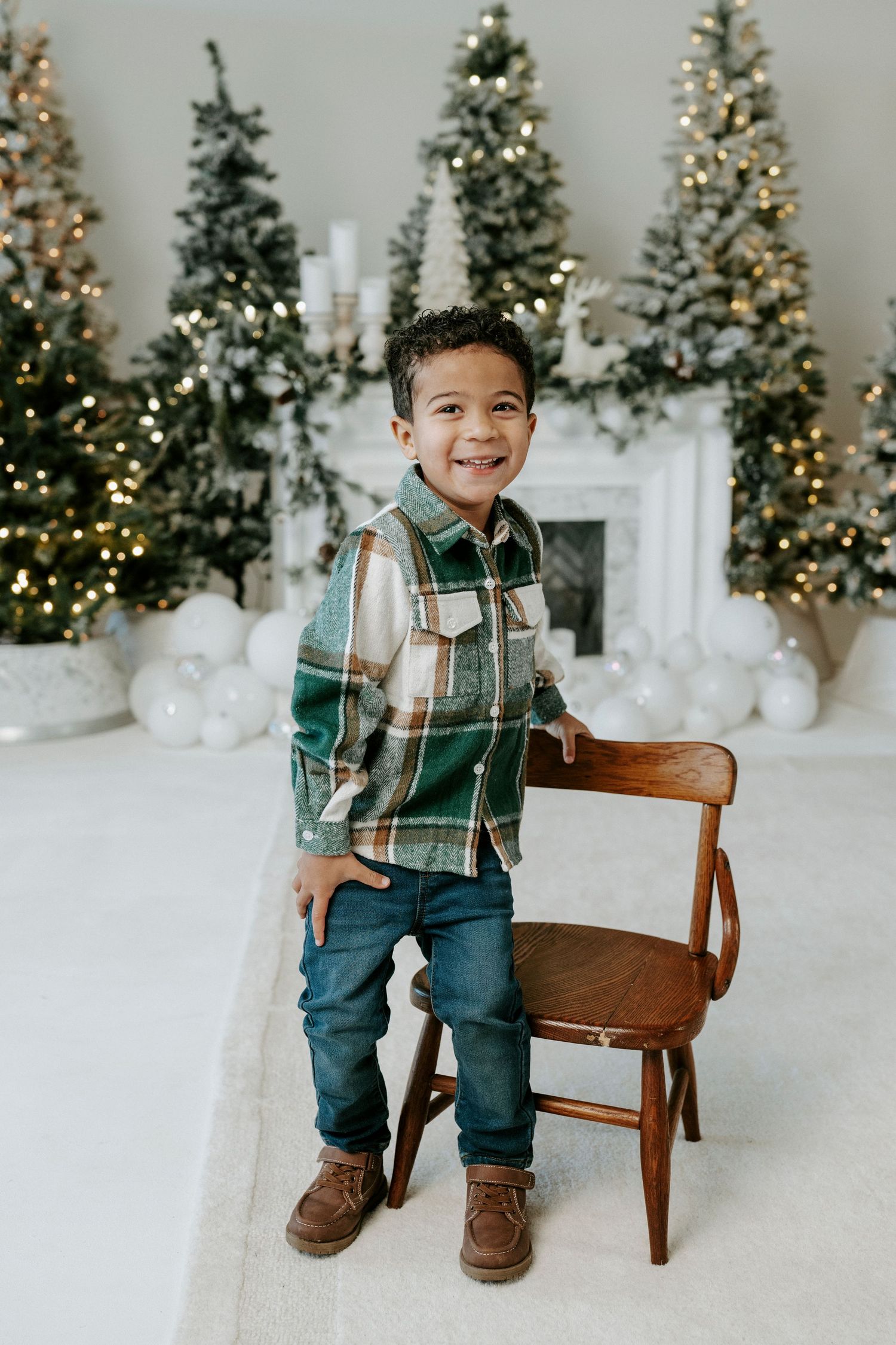 Festive holiday photo session featuring a small child in plaid shirt and jeans sitting on wooden bench with lit Christmas trees.