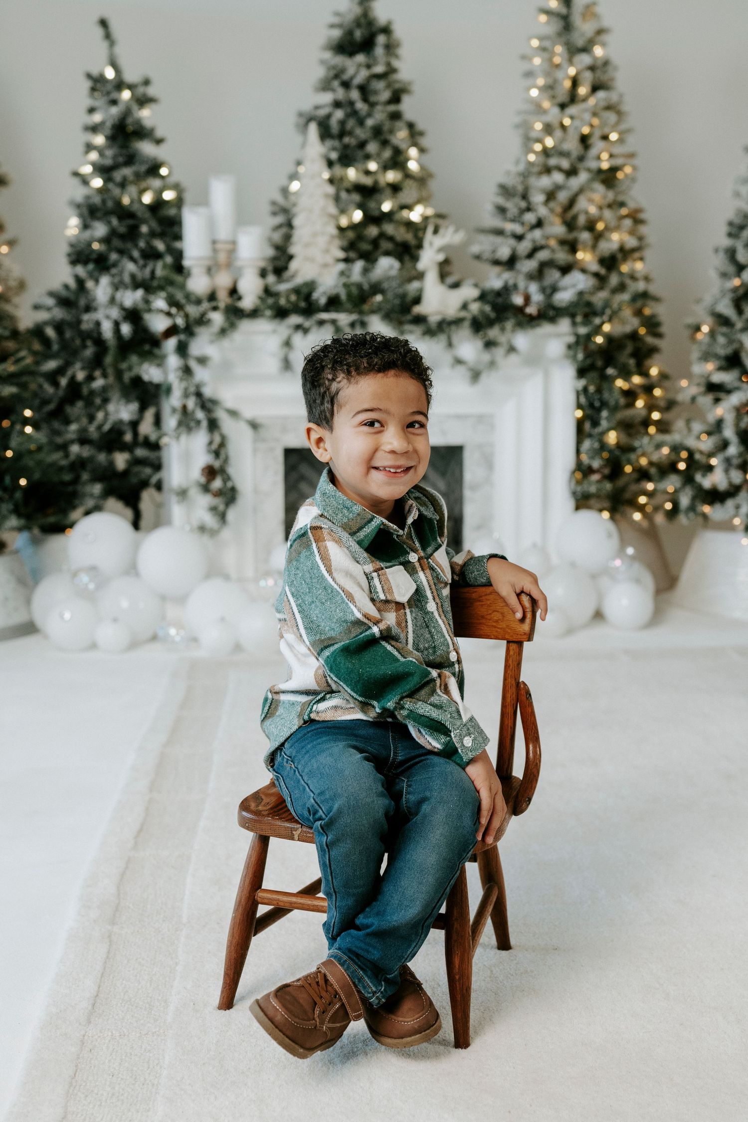 Adorable child in plaid scarf and jeans poses playfully on wooden chair against winter wonderland backdrop with lit trees.