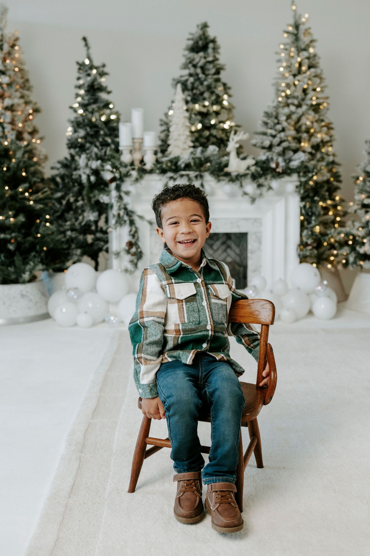 A child sits in a wooden chair wearing plaid and jeans against a backdrop of lit Christmas trees and white decor.