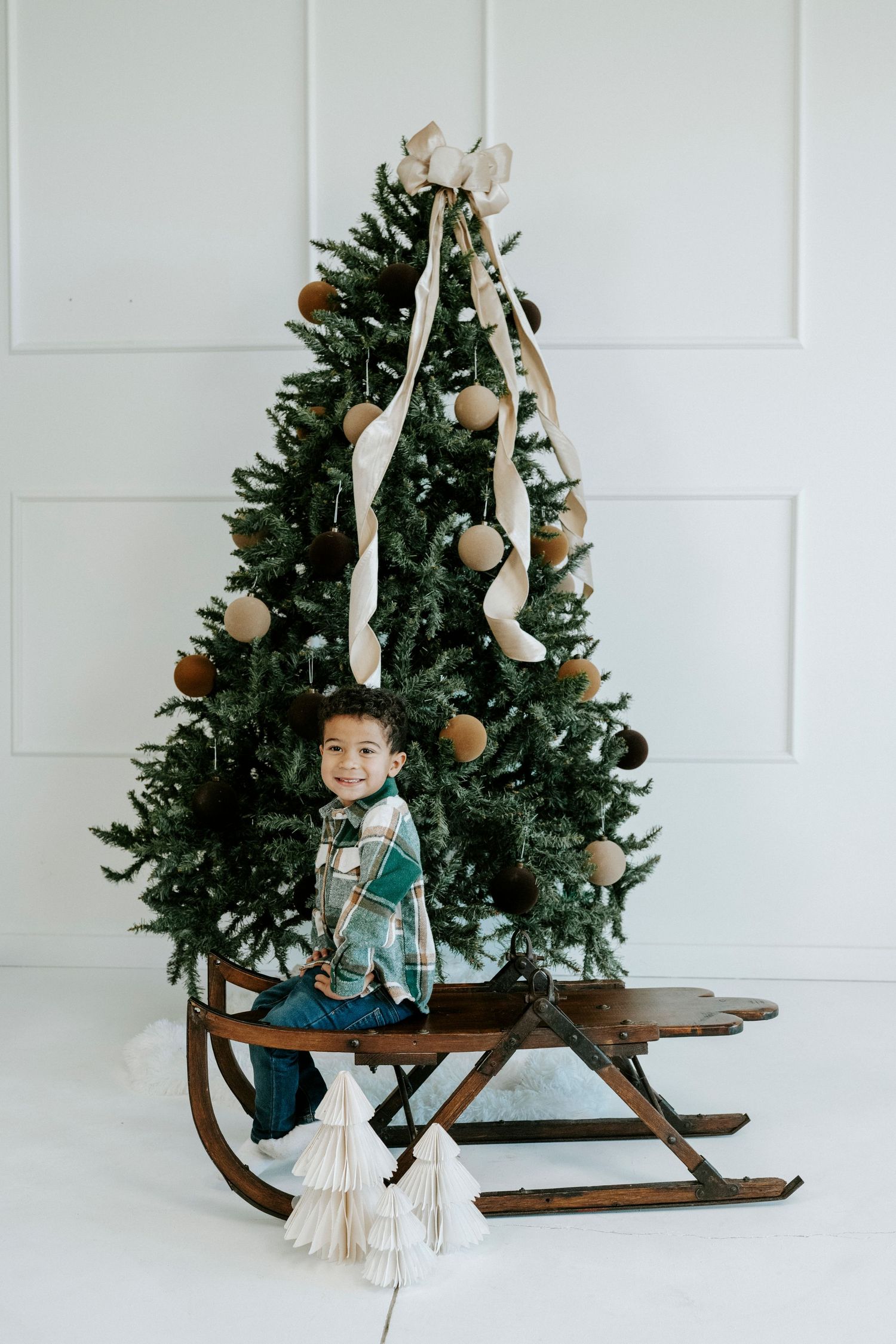 Decorated Christmas tree with vintage wooden sled and rustic ornaments against white wall.