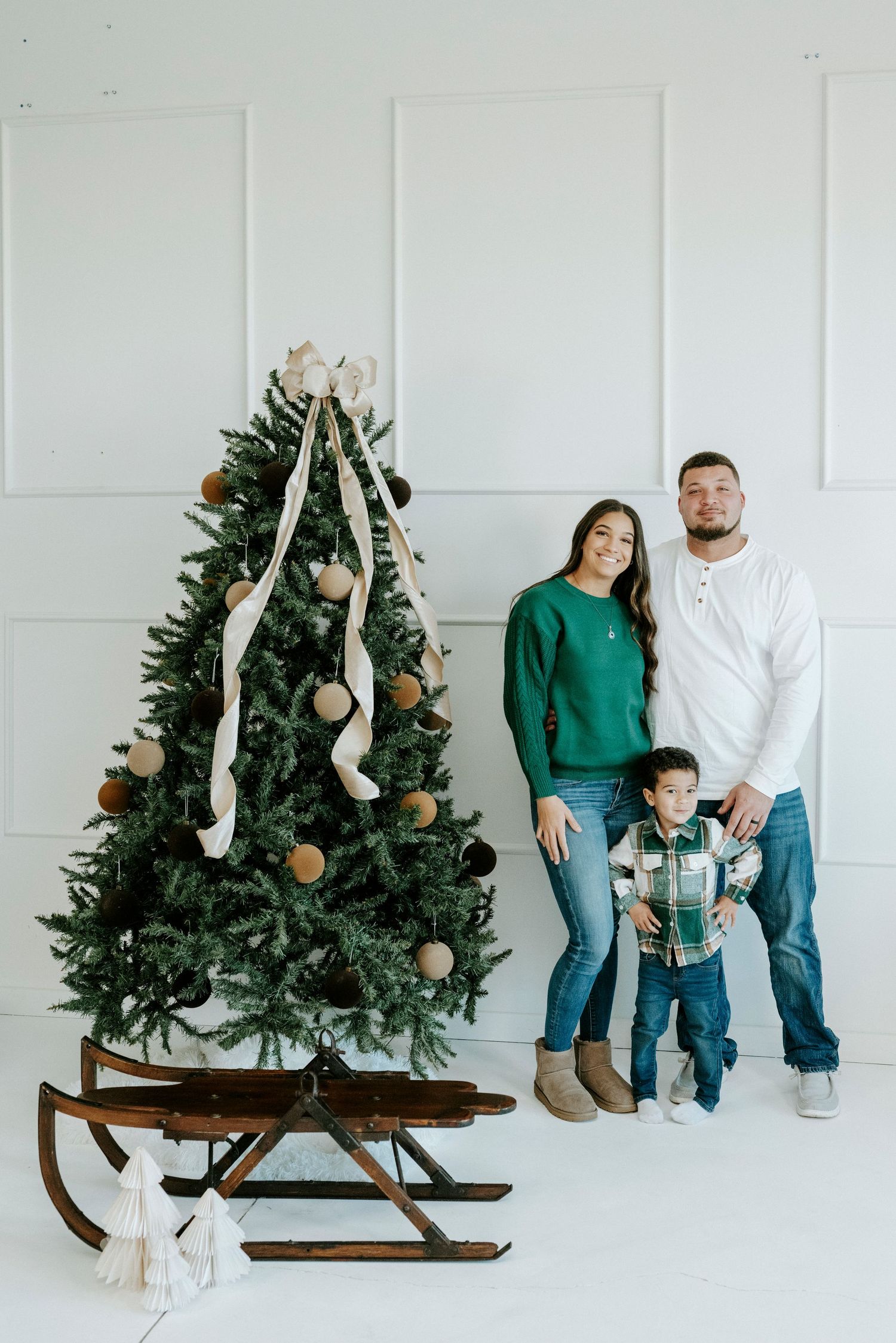 Family poses with vintage wooden sleigh and decorated Christmas tree in minimalist white studio setting.
